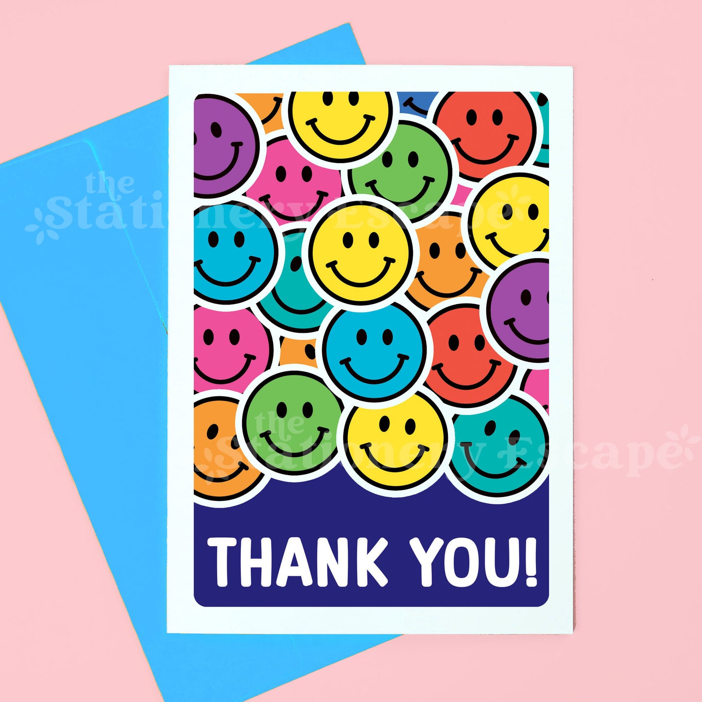 A greeting card with a colorful pattern of overlapping smiley faces and the words 'THANK YOU!' in white on a dark blue band, shown with a bright blue envelope on a pink background.