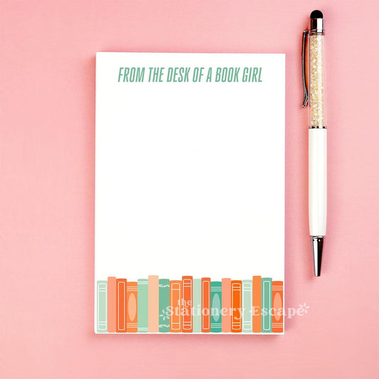 Notepad with 'From the Desk of a Book Girl' text, orange and green book border, and a pen on a pink background