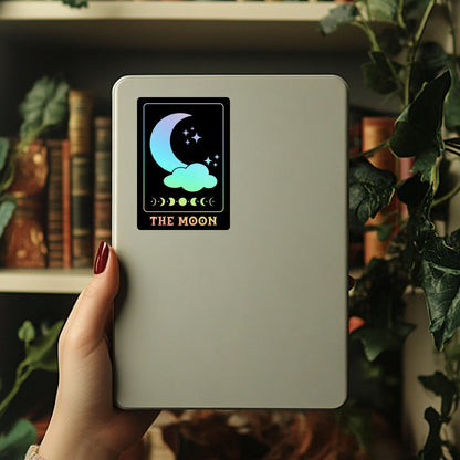 Hand holding a device with a rainbow holographic moon phase tarot card sticker against a blurred bookshelf background