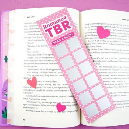 Open book with a pink 'Romance TBR' bookmark and heart stickers on a pink background