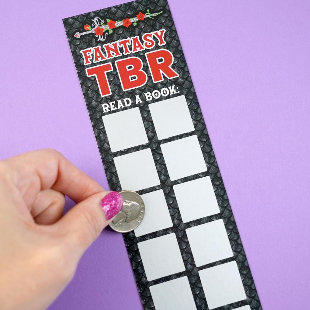Scratch-off bookmark with 'Fantasy TBR' text and a hand placing a coin on it against a purple background