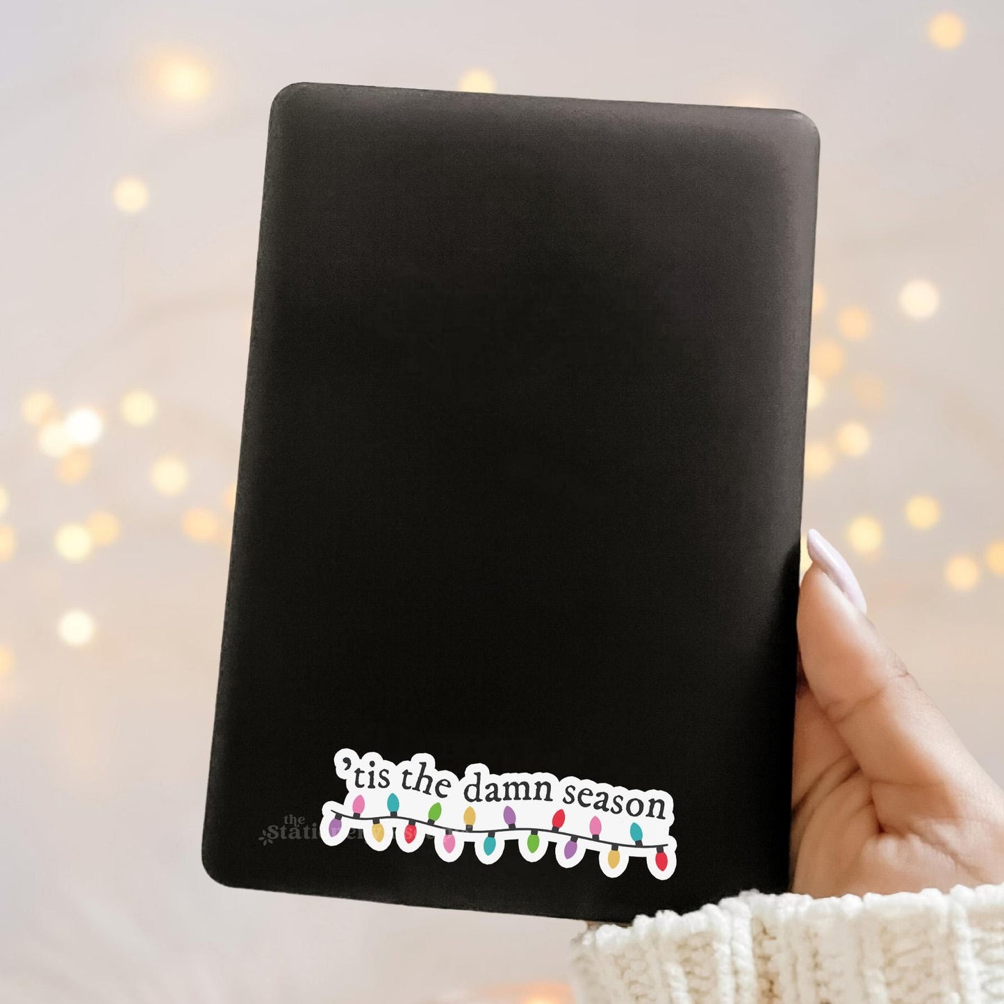 Black Kindle case with 'tis the damn season text and Christmas lights design held by a hand against a blurred light background.