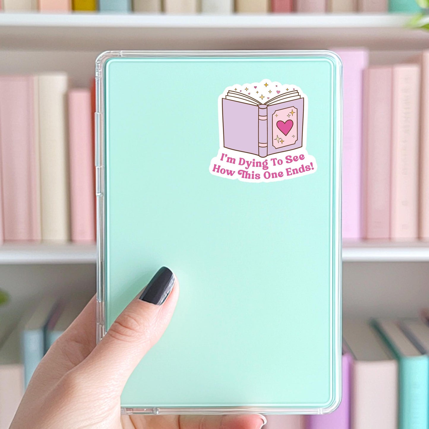 Kindle with a book-themed sticker held by a hand in front of a bookshelf.