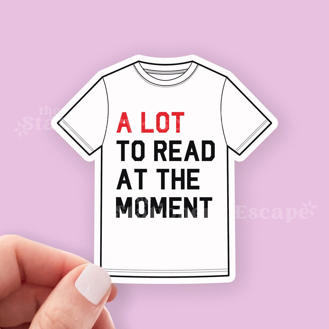 Sticker of a t-shirt with text 'A LOT TO READ AT THE MOMENT' held by a hand on a pink background.