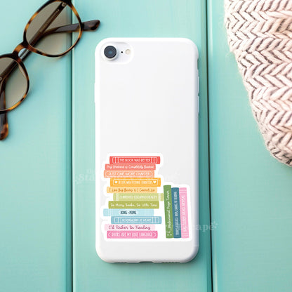 Phone case with colorful book stack design on a light blue surface with sunglasses and a woven bag.