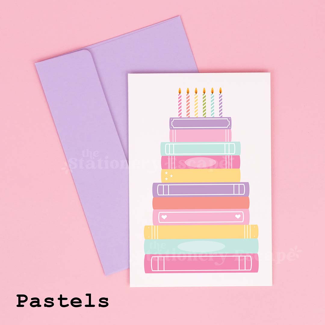 Colorful birthday card featuring a "birthday cake" made from pastel stacked books on a pink background with a lavender envelope