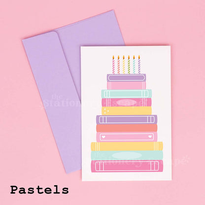 Colorful birthday card featuring a "birthday cake" made from pastel stacked books on a pink background with a lavender envelope