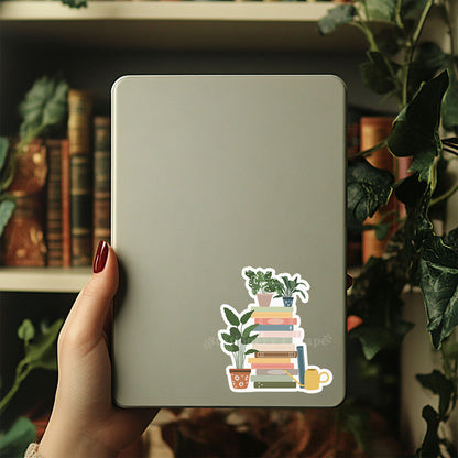 Hand holding a tablet with a sticker of books and plants on a blurred bookshelf background