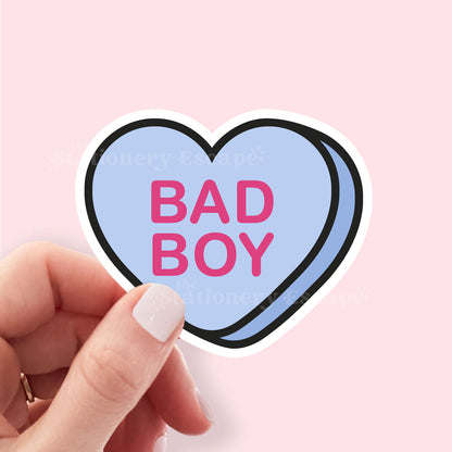 Heart-shaped sticker with 'BAD BOY' text held by a hand against a pink background