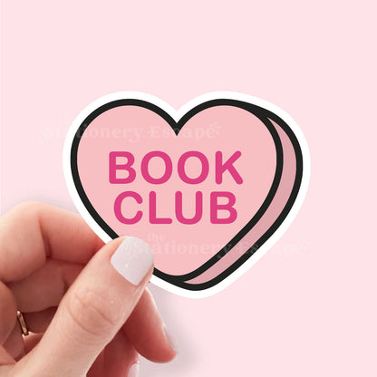 Heart-shaped 'Book Club' sticker held by a hand on a pink background