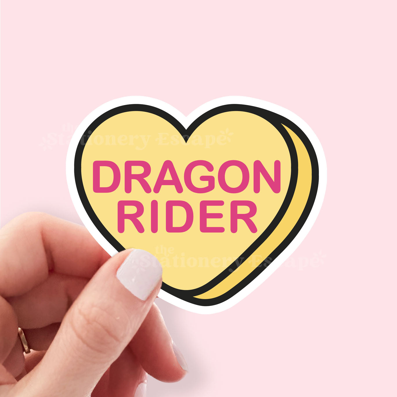 Heart-shaped sticker with 'Dragon Rider' text held by a hand on a pink background