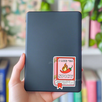 Black Kindle case with a 'I Like 'Em Spicy' sticker held by a hand against a blurred bookshelf background