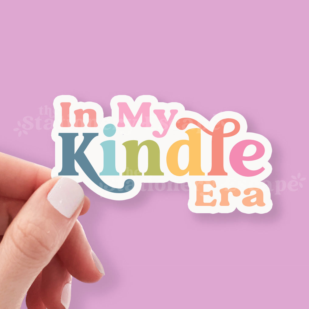 Hand holding a colorful sticker with 'In My Kindle Era' text against a pink background