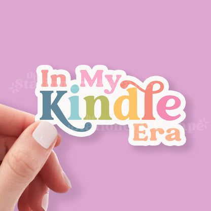 Hand holding a colorful sticker with 'In My Kindle Era' text against a pink background