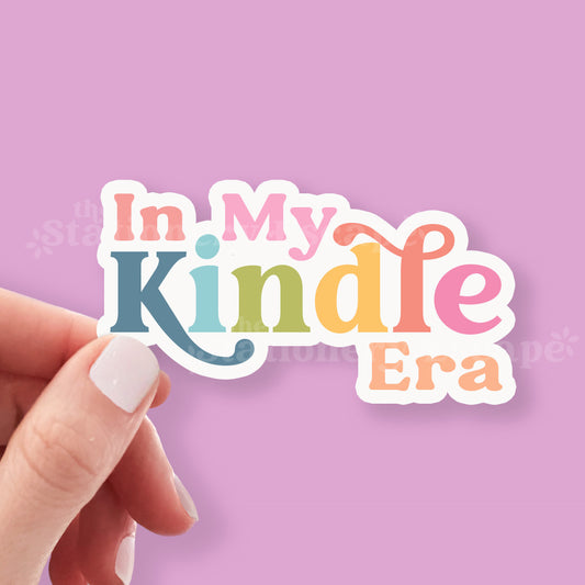 Hand holding a colorful sticker with 'In My Kindle Era' text against a pink background