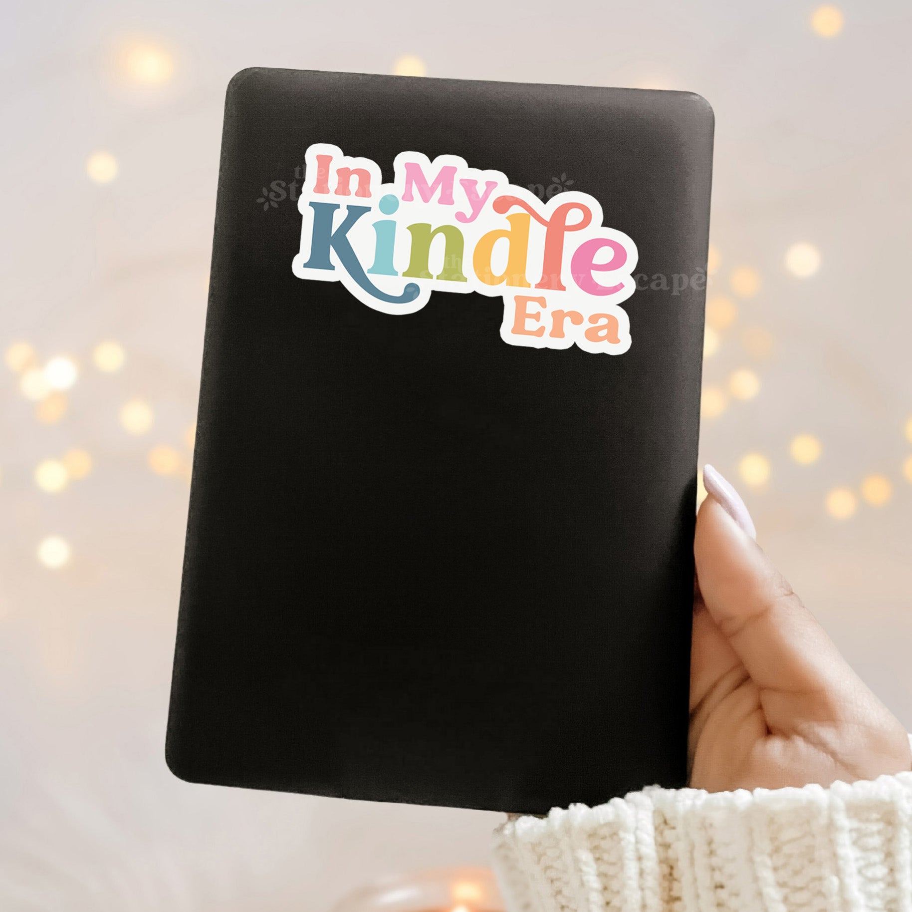 Black Kindle cover with colorful text 'In My Kindle Era' held by a hand against a blurred light background