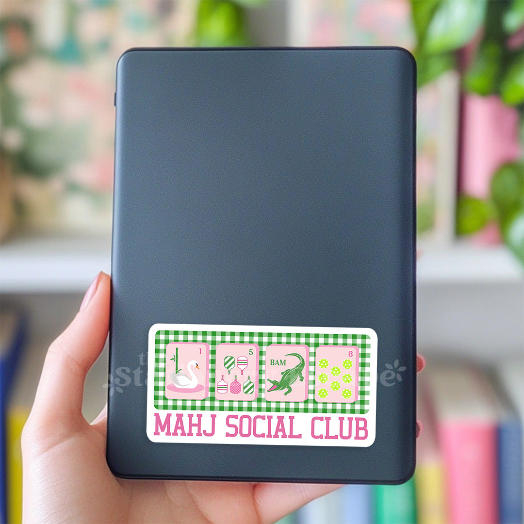 Black tablet with a decorative sticker labeled 'MAHJ SOCIAL CLUB' held by a hand against a blurred indoor background.