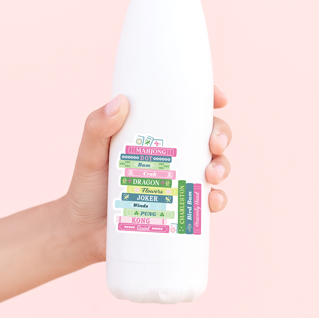 Hand holding a white bottle with colorful mahjong book stack sticker on a pink background
