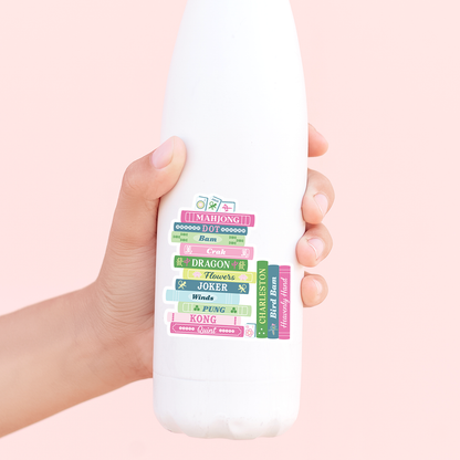 Hand holding a white bottle with colorful mahjong book stack sticker on a pink background