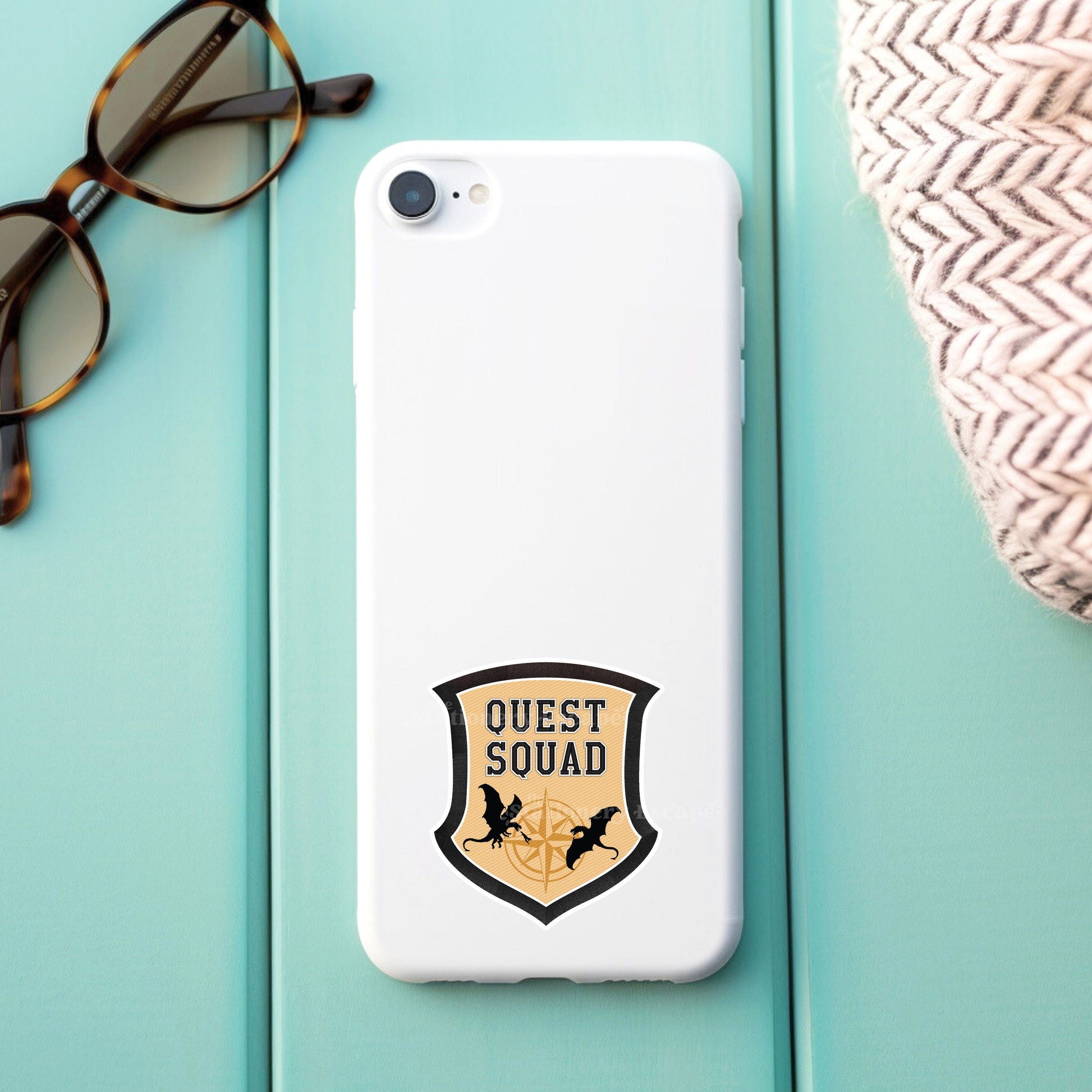 Phone case with 'Quest Squad' sticker design on a light blue surface with sunglasses and a woven bag.