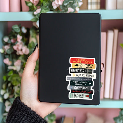Black Kindle case with a romantasy book title sticker held by a hand against a blurred bookshelf background