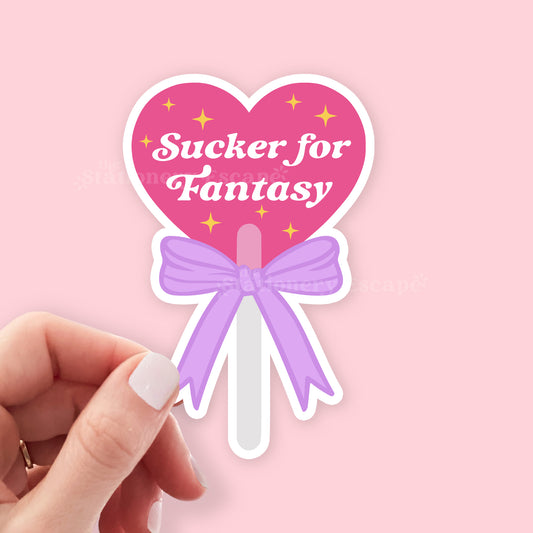 Heart-shaped lollipop sticker with 'Sucker for Fantasy' text on a pink background