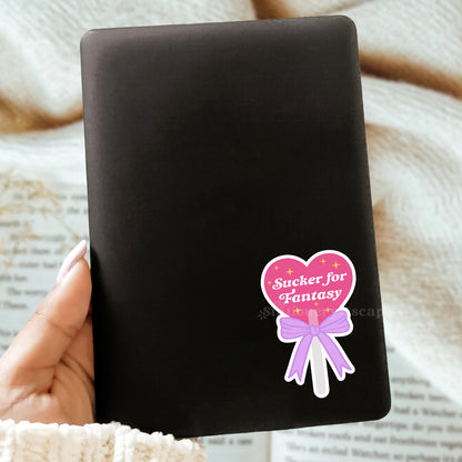 Black Kindle case with a pink and purple sticker featuring a heart lollipop with 'Sucker for Fantasy' text, held by a hand on a textured surface.