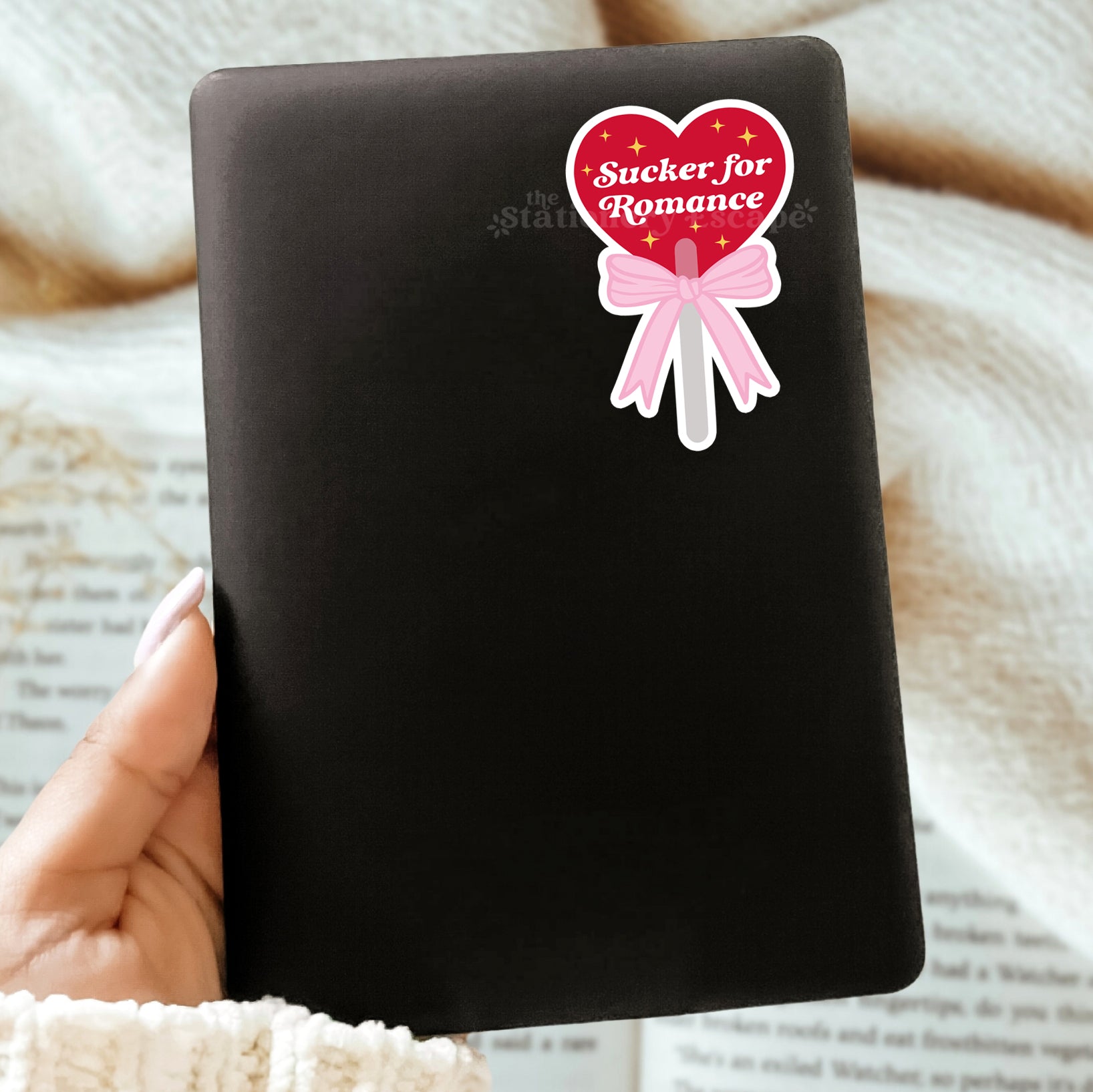 Black Kindle case with a heart-shaped sticker labeled 'Sucker for Romance' held by a hand on a textured surface.