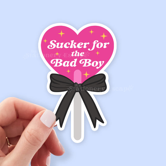 Heart-shaped sticker with 'Sucker for the Bad Boy' text and a black bow, held by a hand against a light blue background.