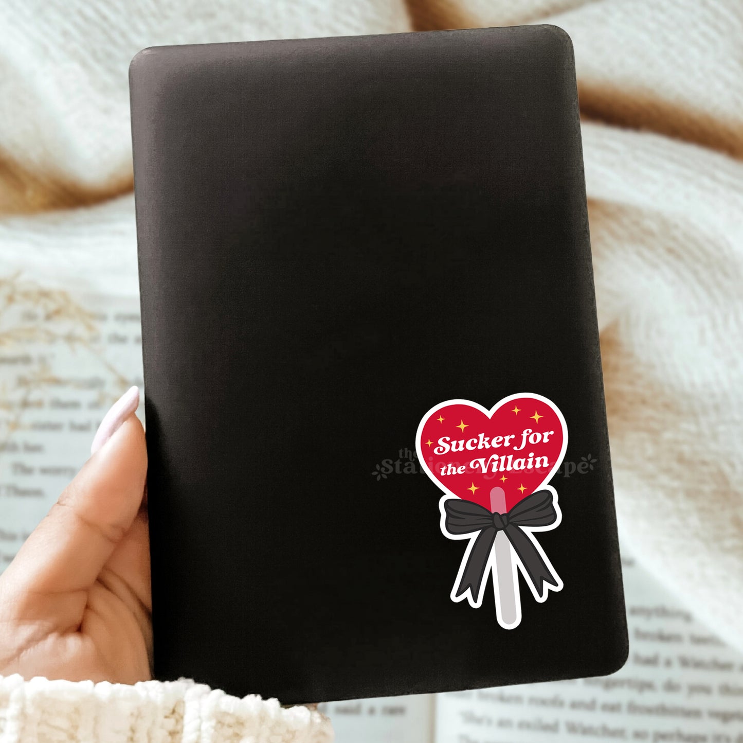 Black Kindle with a heart lollipop sticker with "Sucker for the Villain" text held by a hand on a textured surface