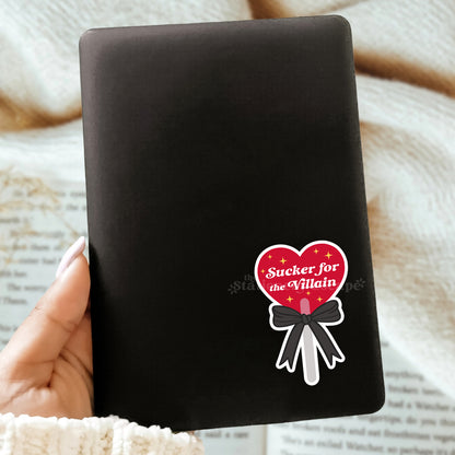 Black Kindle with a heart lollipop sticker with "Sucker for the Villain" text held by a hand on a textured surface
