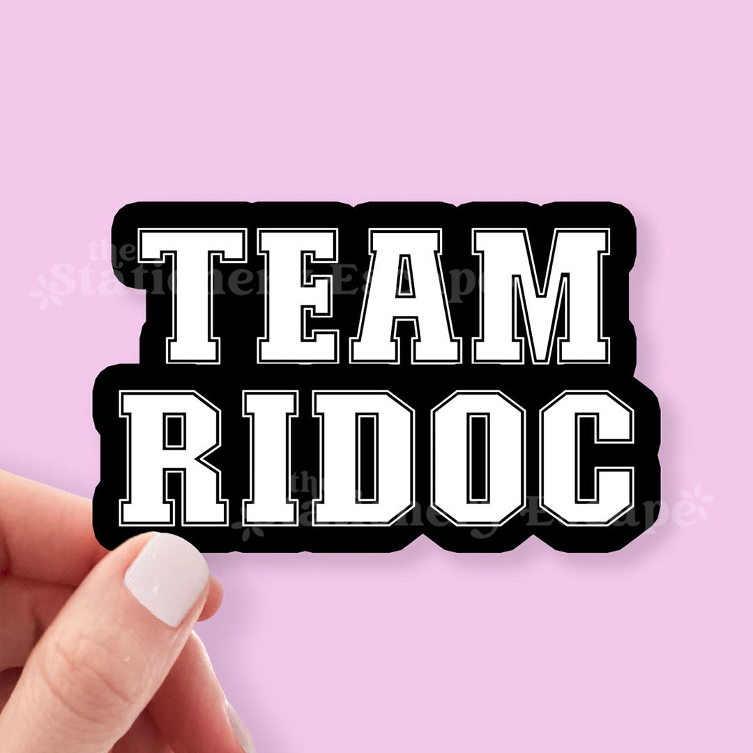 Sticker with 'TEAM RIDOC' text held by a hand against a pink background