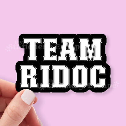 Sticker with 'TEAM RIDOC' text held by a hand against a pink background