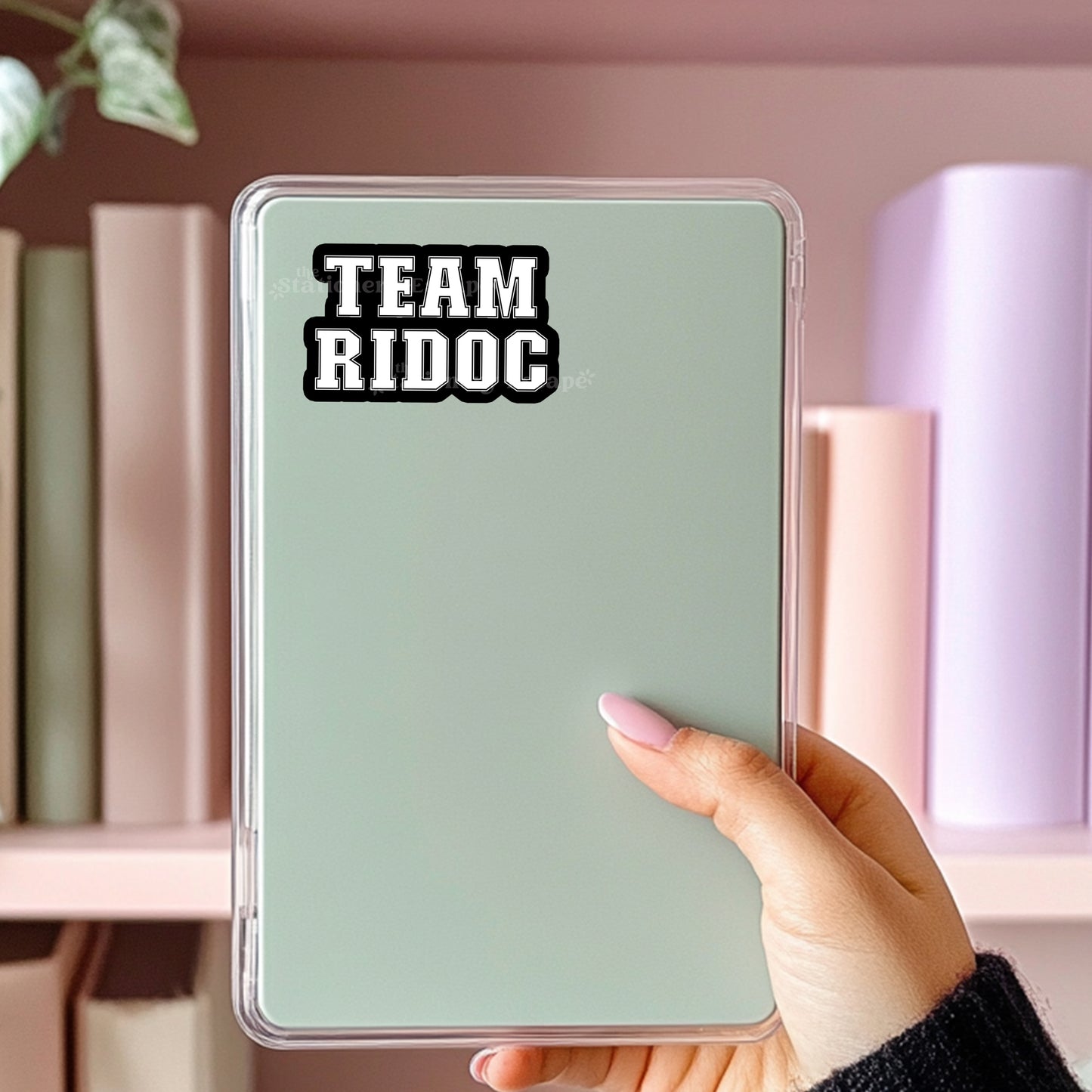 Hand holding a light green Kindle case with 'TEAM RIDOC' sticker against a bookshelf background