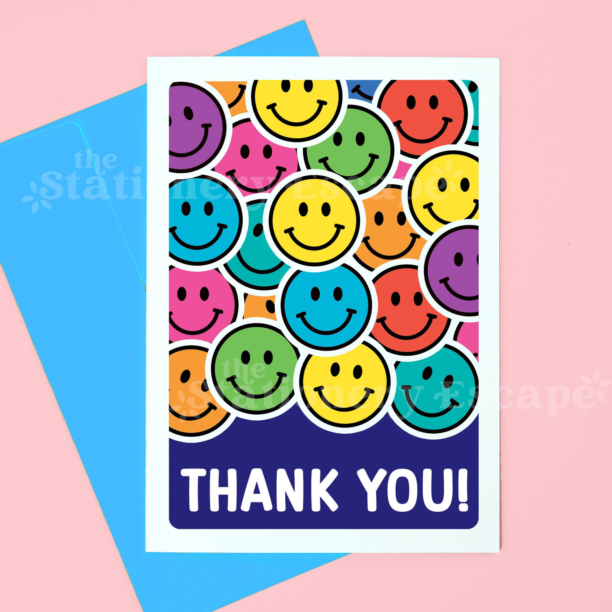A greeting card with a colorful pattern of overlapping smiley faces and the words 'THANK YOU!' in white on a dark blue band, shown with a bright blue envelope on a pink background.