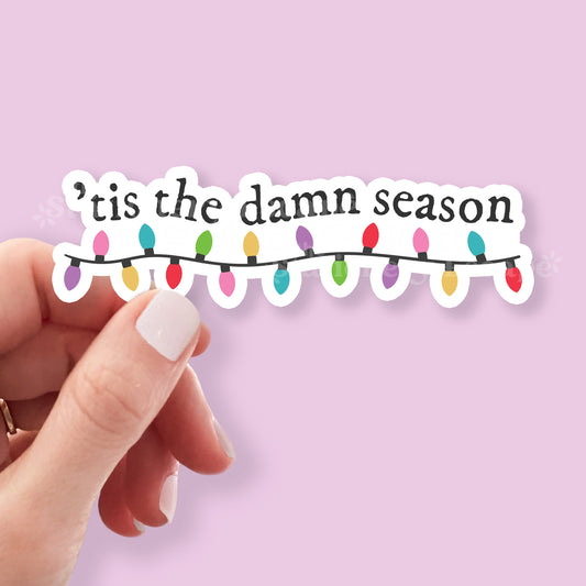 Hand holding a sticker with 'tis the damn season text and Christmas lights on a pink background