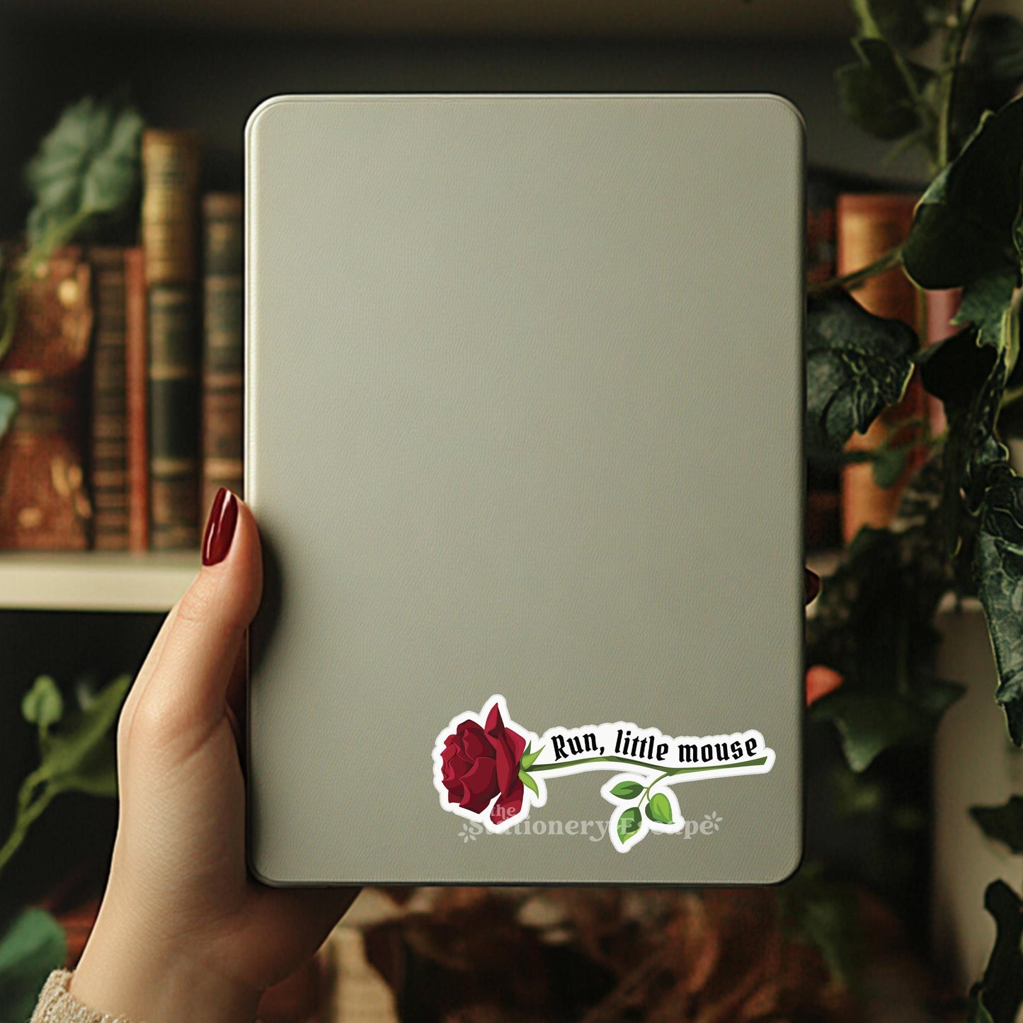Hand holding a light gray notebook with a 'Run, little mouse' sticker on a blurred bookshelf background