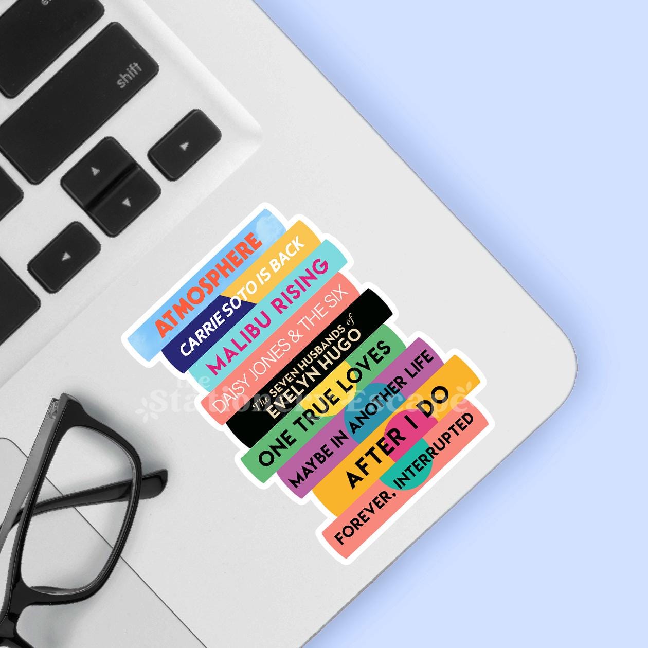 Colorful Taylor Jenkins Reid book title stickers on a laptop keyboard with glasses and a blue background